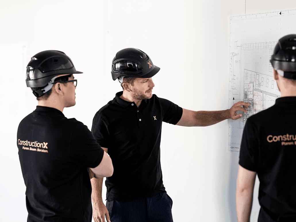 ConstructionX-Team mit Bauhelmen prüft einen Werkplan an der Wand